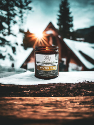 A Winter Sun™ - 9oz. Amber Jar Candle by Begonia & Bench® is placed on a snow-covered wooden surface. In the background, there is a snow-covered rustic cabin surrounded by evergreen trees, with a glowing sun peeking through and casting a warm light. This scented candle creates an ambiance that feels both cozy and serene.