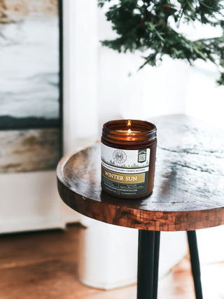 An amber-colored jar labeled "Winter Sun™ - 9oz. Amber Jar Candle - By Begonia & Bench®" sits on a small, round wooden table with metal legs. The fall candle is lit, casting a warm glow. The background features a branch of an evergreen tree and a blurred window, suggesting a cozy indoor winter setting.