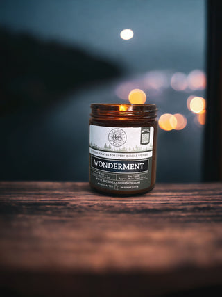 A lit Wonderment™ - 9oz. Amber Jar Candle by Begonia & Bench® is centered on a wooden surface. Behind it, blurred city lights create a bokeh effect against a dark, out-of-focus background. The warm candlelight contrasts with the cool tones of the backdrop, evoking a cozy atmosphere.
