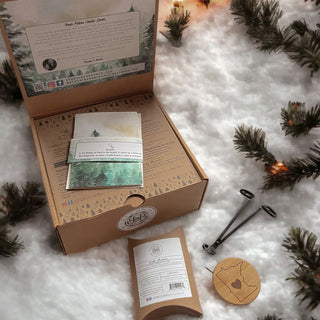 An open cardboard box rests on faux snow, decorated with pine sprigs, and contains the Begonia & Bench Winter Solstice Gift Box featuring an artisan candle, a booklet of forest imagery, and a care card. Next to it is a wick trimmer and a charming wooden Michigan plaque. The lid displays instructions and company details.