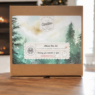 A brown cardboard box labeled with "Candles" and featuring a beautiful design of green pine trees and mist showcases the words "Artisan Candles" and "Wishing you warmth & light." This Begonia & Bench Winter Solstice Gift Box radiates coziness in a setting with a softly blurred background, warm-lit interior, and an inviting fireplace.