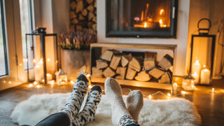 Two pairs of feet in cozy socks rest on a fluffy white rug in front of a fireplace. The scene is warmly lit by numerous candles and the glow from the fire. Stacked firewood is visible next to the fireplace, and lanterns and decorative elements enhance the cozy atmosphere.