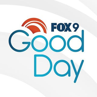 The image shows the "FOX 9 Good Day" logo. An orange semi-circle with three arcs resembling a sun is above the word "Good," which is written in large, blue letters with "Day" underneath in matching blue text. "FOX 9" is in bold, navy blue text to the upper right of the arcs. The background is white with light grey curves.