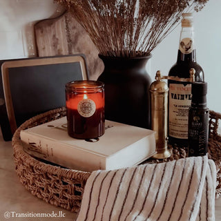 A cozy kitchen scene showcases a woven tray containing a French Oak™ - 12oz Finely Crafted Scented Candle By Begonia & Bench®, a stack of books, and a striped cloth. In the background, dried flowers in a dark vase, vanilla extract, and a pepper grinder rest against a French oak cutting board. Text reads "@Transitionmode.llc.