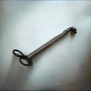 A pair of Wick Trimmers with scissor-like handles and a flat, elongated body is lying on a smooth, light-colored surface beside a scented candle. The trimmers feature a small catchment tray near the cutting end to hold the trimmed wick pieces.