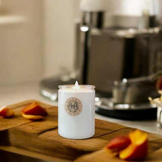 A lit Pesche Dolce™ - 12oz Finely Crafted Scented Candle by Begonia & Bench® in a glass jar sits on a wooden cutting board, surrounded by juicy peaches in a kitchen setting. The jar, adorned with an emblem, complements the Italian Sweet Peach theme. A blurred background featuring kitchen appliances like a mixer enhances the cozy, homely atmosphere.