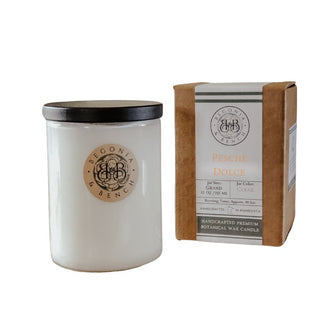 A Pesche Dolce™ 12oz finely crafted scented candle by Begonia & Bench® in a white glass jar with a black lid is placed next to a brown cardboard box. Both items feature matching labels displaying the brand "BEGONIA & BENCH" and the scent "Italian Sweet Peach." This handcrafted premium botanical wax candle from Minnesota embodies the essence of succulent peaches.