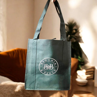 In a sunlit room, someone holds up a Begonia & Bench Reusable Shopping Tote, dark green in color with the name "Begonia & Bench" printed in white on the front. The setting includes a brown couch against a white wall and hints of blurred greenery, creating a cozy and inviting atmosphere ideal for autumn getaways or visits to the farmer's market.