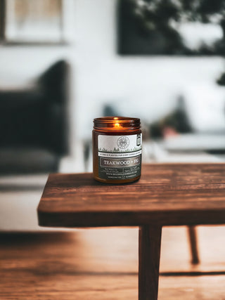 A lit Teakwood & Fig™ - 9oz. Amber Jar Candle by Begonia & Bench® sits on a wooden table. The background features a blurred view of a sofa, a cushion, and some greenery, suggesting a cozy, well-decorated living space. The scented candle exudes warmth and a serene atmosphere.