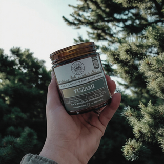 A hand holds a brown glass jar candle labeled "Yuzami™ - 9oz. Amber Jar Candle - By Begonia & Bench®" in front of lush evergreen trees. The label highlights eco-friendly features, including tree planting initiatives. Infused with hints of yuzu, the natural lighting enhances the serene and earthy outdoor ambiance.