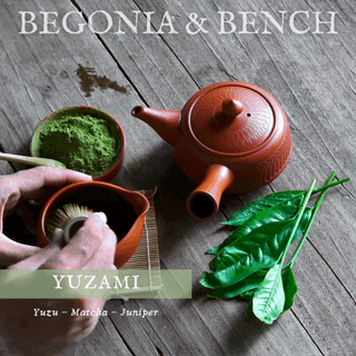 A Japanese-style tea display features a terracotta teapot, a bowl with vibrant matcha being whisked, and green tea leaves arranged on a wooden surface. "Begonia & Bench®" is prominently featured at the top, while "Yuzami: Yuzu - Matcha - Juniper," reminiscent of the 9oz. Amber Jar Candle by Begonia & Bench®, is elegantly scripted at the bottom.