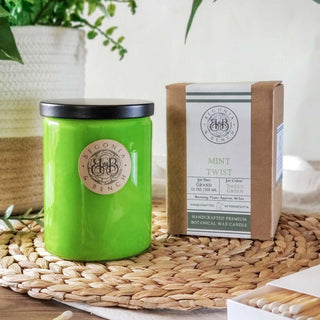 A bright green scented candle in a glass jar with a black lid, labeled "Mint Twist™ - 12oz Finely Crafted Scented Candle By Begonia & Bench®." Next to it is a cardboard box with matching branding. The items are placed on a woven placemat with a green plant in a pot partially visible in the background.