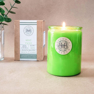 A lit green candle in a glass jar sits on a light brown surface. The jar has a label with "Begonia & Bench®" on it. Behind the scented candle is a box with the same branding, displaying "Mint Twist™ - 12oz Finely Crafted Scented Candle By Begonia & Bench®" and various details. A sprig of green leaves in a clear glass vase is partially visible on the left.