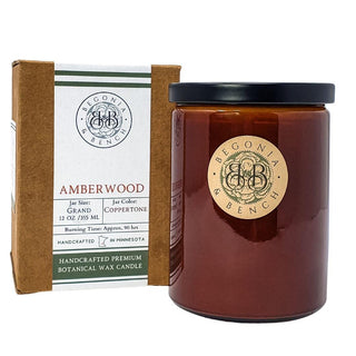 A 12 oz jar of Amberwood - Finely Crafted Scented Candle by Begonia & Bench® in a copper-toned finish stands next to its brown box packaging. The packaging features product details: jar color, burn time (approx. 90 hours), and a "Handcrafted in Minnesota" label.