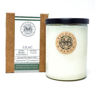 A white scented candle in a transparent glass jar with a black lid stands next to a brown cardboard box. The jar has a circular wooden label with the text "Begonia & Bench®." The box displays details including "Lilac," "12 oz," "Burning Time: Approx. 90 hrs," and "Handcrafted in Minnesota, featuring the product name 'Lilac - 12oz Finely Crafted Scented Candle By Begonia & Bench®.'