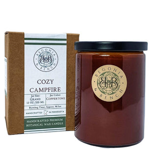 A "Cozy Campfire™ - 12oz Finely Crafted Scented Candle By Begonia & Bench®" is displayed. The scented candle, housed in a glossy amber jar with a black lid, sits beside its brown and white packaging. The label on the jar and packaging features the brand logo and candle details.