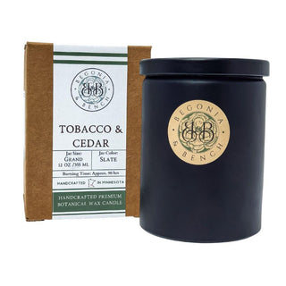 A 12oz finely crafted scented candle by Begonia & Bench®, named Tobacco & Cedar™, with a brown cardboard box beside it. The box labels the fall candle as handcrafted in Minnesota with an approximate burning time of 90 hours. The candle is housed in a sleek, dark-colored jar adorned with a round, beige label featuring the Begonia & Bench® logo.