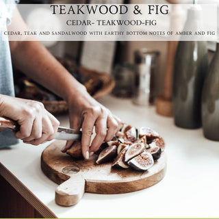 A person is slicing figs on a round wooden cutting board. The text "TEAKWOOD & FIG, CEDAR - TEAKWOOD - FIG" is at the top, while "CEDAR, TEAK AND SANDALWOOD WITH EARTHY BOTTOM NOTES OF AMBER AND FIG" lies below it. Various kitchen items and ingredients are visible in the background, enhancing the 12oz Teakwood & Fig™ Finely Crafted Scented Candle by Begonia & Bench®.