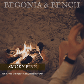 A person is sitting by a cozy small campfire, resting their hand on their head. The text above reads "Begonia & Bench®" and the text below the fire says "Smoky Pine™ - 9oz. Amber Jar Candle." At the bottom, descriptive text indicates "Pinewood embers-Marshmallow-Oak." The scene suggests a warm, rustic ambiance.