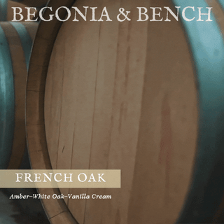 A close-up image of a French oak barrel with the text "Begonia & Bench®" at the top. Below, there's a label that reads "French Oak™ - 9oz. Amber Jar Candle," which describes the warm blend of amber, white oak, and vanilla cream. The background features blurred, similar barrels, contributing to a rustic and aged ambiance reminiscent of the cozy warmth provided by this fall-inspired candle.