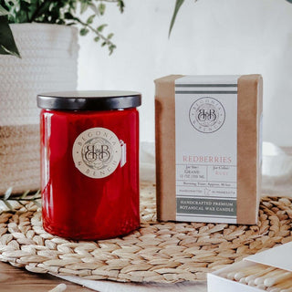 A red jar candle with a black lid labeled "Begonia Essence" sits next to a wrapped box labeled "Redberries™ - 12oz Finely Crafted Scented Candle By Begonia & Bench®" on a woven mat. Behind them, out of focus, are a white pot with a plant and a stack of books. The setting is bright and inviting, suggesting a cozy atmosphere perfect for lighting your favorite fall candle.