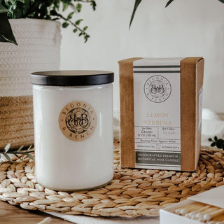 A 12oz finely crafted Lemon Verbena scented candle by Begonia & Bench® sits on a woven mat next to its tan-colored packaging. The white botanical candle, housed in a glass jar with a black lid, and its box both feature the same elegant branding. Houseplants and a cozy, neutral background add to the perfect fall ambiance.