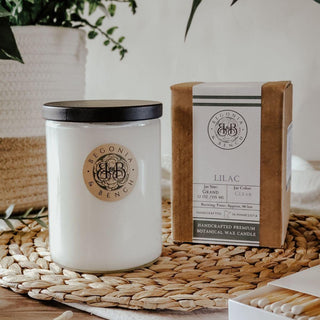 A Lilac - 12oz Finely Crafted Scented Candle By Begonia & Bench® in a clear jar with a black lid and a round "Begonia & Bench" label sits next to its brown and white packaging on a wicker placemat. The label on the box indicates the scent is "Lilac," and the fall candle is handcrafted from premium botanical wax. Matches are visible in the foreground.