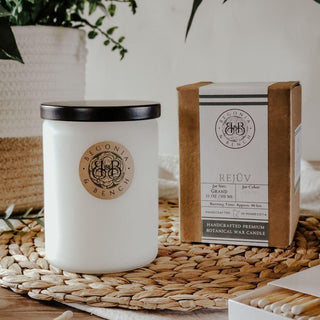 A white cylindrical scented candle with a black lid is placed on a woven mat. The candle label reads "Rejüv™ - 12oz Finely Crafted Scented Candle By Begonia & Bench®." Beside it sits a brown box with similar branding, labeled "Rejuv." Various decorative items and plant leaves in the background create a cozy and inviting scene.