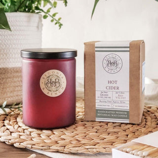 A red candle jar with a black lid labeled "Begonia & Bench" sits on a woven mat. Beside it, a matching brown box labeled "Hot Cider™ - 12oz Finely Crafted Scented Candle By Begonia & Bench®" provides details about the fall-scented candle inside, including the handcrafted premium botanical wax, scent notes, burn time, and size. A potted plant is in the background.