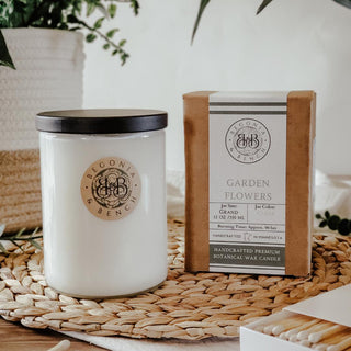 On a wicker mat sits the Garden Flowers™ 12oz Finely Crafted Scented Candle By Begonia & Bench® in a white glass jar with a black lid. Next to it, the candle’s packaging displays "Garden Flowers." In the background, there’s a white pot with a plant and a book. A matchbox is partially visible in the lower right corner.