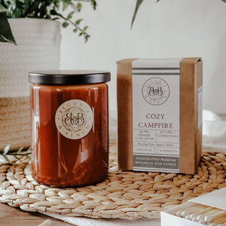 A cozy setup features a brown jar candle with a black lid labeled "Begonia & Bench." Next to it is a brown cardboard box labeled "Cozy Campfire™ - 12oz Finely Crafted Scented Candle By Begonia & Bench®." Both are placed on a woven mat, with a potted plant and other small decor items in the background.