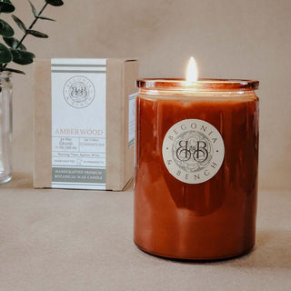 A lit Amberwood scented candle in a 12oz glass jar bearing the "Begonia & Bench®" label sits on a table. Behind it, there is a brown box with a similar label and "Amberwood - 12oz Finely Crafted Scented Candle By Begonia & Bench®" written on it. The background is neutral beige, and a small branch with green leaves is partially visible on the left.