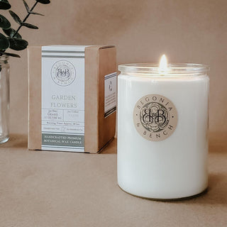 A lit "Garden Flowers™ - 12oz Finely Crafted Scented Candle By Begonia & Bench®" sits on a beige surface. The scented candle is in a glass container with white wax. Beside it is a brown box with the product label, detailing the candle's specifications. A green leafy plant is partially visible in the background.