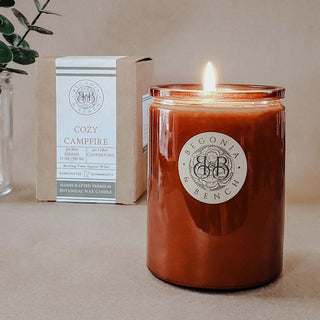 A lit, amber-colored glass candle labeled "Cozy Campfire™ - 12oz Finely Crafted Scented Candle By Begonia & Bench®" sits on a beige surface. Behind it is a box with the same branding, indicating it is handcrafted from premium botanical wax. Some greens are partially visible on the left side.