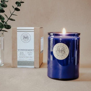 A stylish blue jar candle from the label "Begonia & Bench®" illuminates the scene, sitting next to a matching box labeled "Wonderment™ - 12oz Finely Crafted Scented Candle." This box highlights key features of the scented candle, such as its 12-ounce size and handcrafted botanical wax composition. Delicate greenery peeks in from the left background.