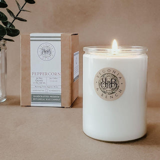 A lit Peppercorn - 12oz Finely Crafted Scented Candle by Begonia & Bench® in a clear glass jar is placed on a beige surface. To the left is the candle’s brown box labeled "Peppercorn." Green leaves in a glass vase adorn the background, set against a neutral brown backdrop.