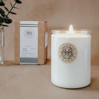 A lit white scented candle in a glass jar labeled "Lilac - 12oz Finely Crafted Scented Candle By Begonia & Bench®" sits on a wooden surface. Beside it, a brown cardboard box marked "Lilac" offers detailed information about the fall candle. A sprig of greenery in a glass jar adds to the cozy ambiance.