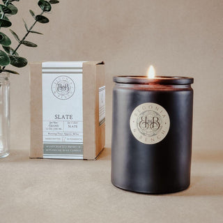 A lit scented candle in a dark, matte container beside its packaging on a beige background. The candle features a beige label with the "Begonia & Bench®" logo. The box beside it is labeled "Slate™ - 12oz Finely Crafted Scented Candle By Begonia & Bench®" and details its net weight and burning time. A sprig of greenery in a clear vase sits to the left.