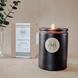 A lit Tobacco & Cedar™ 12oz scented candle by Begonia & Bench® in a matte black jar sits on a beige surface. The label on the jar reads "Begonia & Bench®." Behind the candle, there is a decorative cardboard box featuring the same Begonia & Bench® logo and labeled "Tobacco & Cedar," complete with scent descriptors. Green foliage is partially visible on the left.