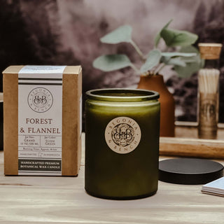 A Forest & Flannel™ 12oz finely crafted scented candle by Begonia & Bench® in a matte green glass jar rests on a wooden surface. Next to it is a brown cardboard box with white labeling that reads "Forest & Flannel." In the background are green leaves in a vase and a glass jar with long wooden matches.