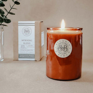 A finely crafted 12oz scented candle from Begonia & Bench®, labeled "Morning Blend™," is lit and placed on a light brown surface in a glass container. Behind it, a box bearing the same label is visible. The background features a clear vase with green foliage on the left, creating an ideal ambiance for fall.