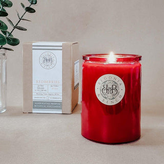 A lit red scented candle in a transparent glass jar with a "Begonia & Bench" label sits on a light brown surface. To its left, there is a boxed candle labeled "Redberries™ - 12oz Finely Crafted Scented Candle By Begonia & Bench®" with details about its size and scent. A sprig of green leaves in a glass vase is partially visible on the left side.