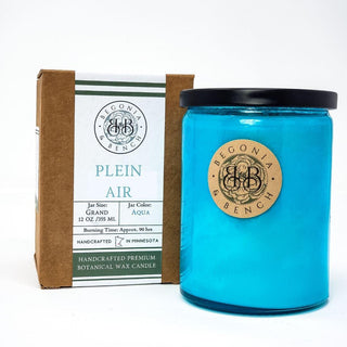 The Plein Air™ - 12oz Finely Crafted Scented Candle by Begonia & Bench® is a botanical wax candle that comes in an aqua blue jar with a black lid and a wooden label. The packaging indicates the candle has a 12 oz (355 ml) capacity, burns for about 90 hours, and is handcrafted in Minnesota. Brown and white packaging can be seen behind the jar.