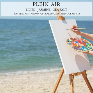 A painter's hand holds a paintbrush and a palette filled with colorful paint, working on a blank canvas placed on an easel set on a sandy beach. The ocean waves are gently visible in the background. Above, text reads "Plein Air™ - Lilies - Jasmine - Sea Salt," evoking an elegant aroma reminiscent of the 12oz finely crafted scented candle by Begonia & Bench®.