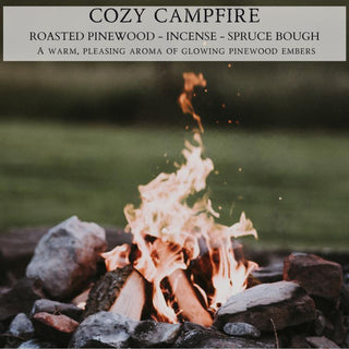 A campfire burns brightly with visible flames amongst stones in a grassy outdoor area. Above, a text box reads: "COZY CAMPFIRE™ - 12oz FINELY CRAFTED SCENTED CANDLE BY BEGONIA & BENCH®: ROASTED PINEWOOD - INCENSE - SPRUCE BOUGH. A warm, pleasing aroma of glowing pinewood embers." Rocks encircle the fire, and a blurred green background is visible.