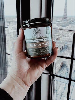A hand holding a lit French Oak™ - 9oz. Amber Jar Candle by Begonia & Bench®. The scented candle, nestled in an amber glass jar with a silver lid, casts a warm glow. Through the window, Paris unfolds with the Eiffel Tower prominently visible in the distance.