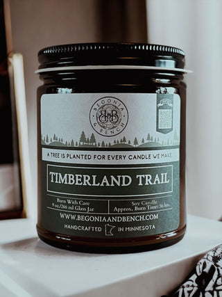 An amber jar with a dark brown hue and a black lid houses the scented candle "Timberland Trail™ - 9oz. Amber Jar Candle," made by Begonia & Bench®. The label features a green and white design, stating: "Timberland Trail," "A tree is planted for every candle we make," alongside details about burning, ingredients, and the website www.begoniaandbench.com.