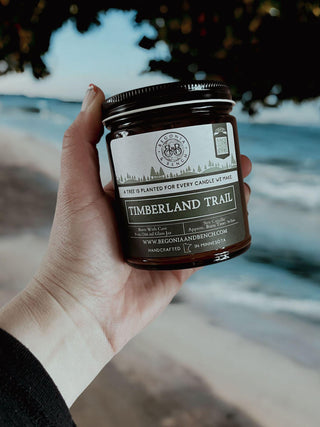 A hand holding a Timberland Trail™ 9oz. Amber Jar Candle by Begonia & Bench®. The label reads: "A tree is planted for every candle we make," and “Handcrafted in Minnesota.” In the background, there's a beach with waves and trees, creating an ideal fall scented candle scene.