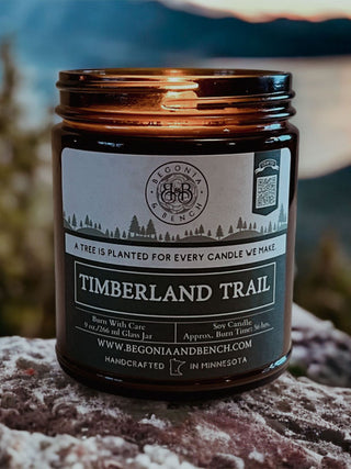 A dark amber glass jar candle labeled "Timberland Trail™ - 9oz. Amber Jar Candle - By Begonia & Bench®." The label mentions it is a soy scented candle with 9 oz / 266 ml of wax, handcrafted in Minnesota. The jar rests outdoors on a stone surface, with blurred greenery and a body of water in the background.
