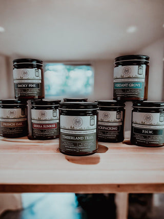 A collection of seven fall candles in dark jars with labels is displayed on a wooden surface. The scented candles are titled Smoky Pine, Sierra Sunrise, Verdant Grove, French Oak, Backpacking, P.N.W., and Timberland Trail™ - 9oz. Amber Jar Candle - By Begonia & Bench® which is centered. The background is softly blurred with natural light.
